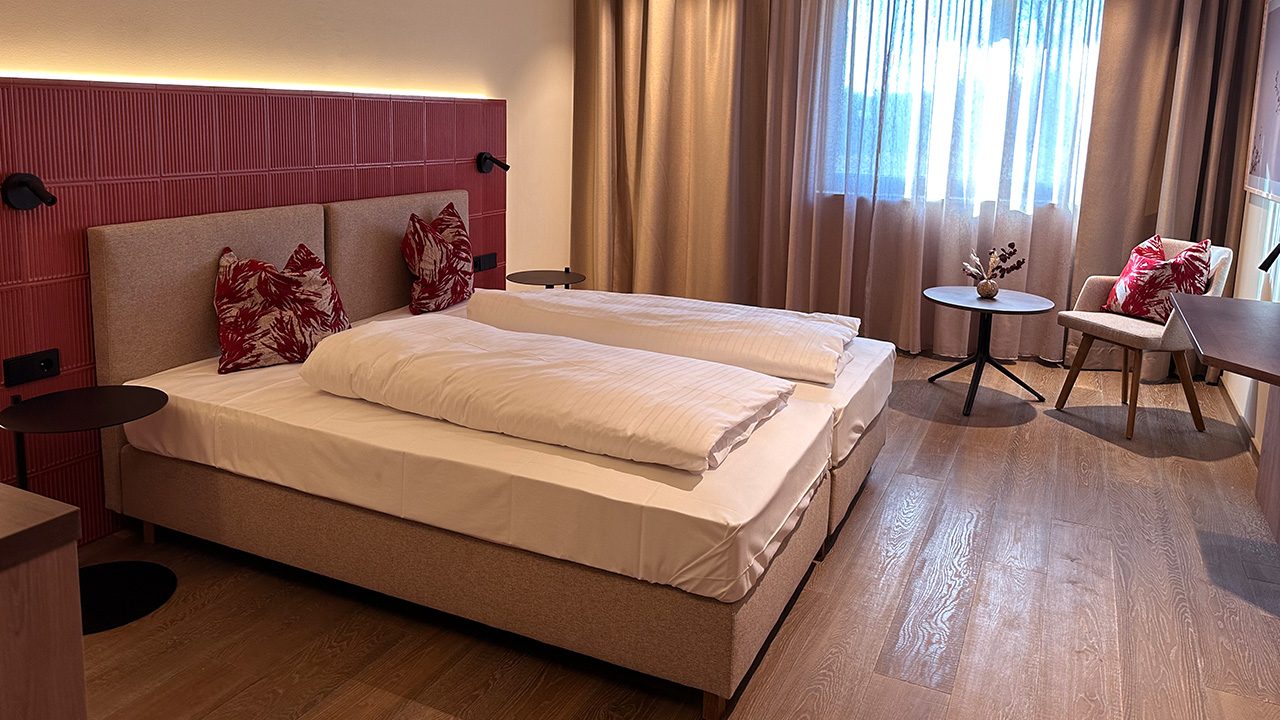 Doppelzimmer mit 20 m²: Doppelbett vor einem rot glasierten Kachel-Kopfteil, daneben ein kleiner Tisch mit Sessel am Fenster